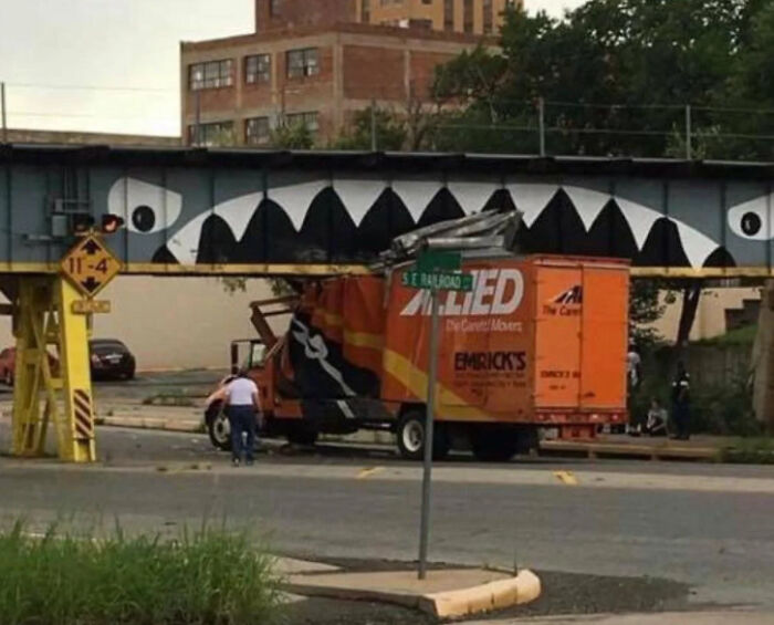 A moving truck stuck under a low bridge with painted teeth, showcasing a humorous parking fail on a city street.