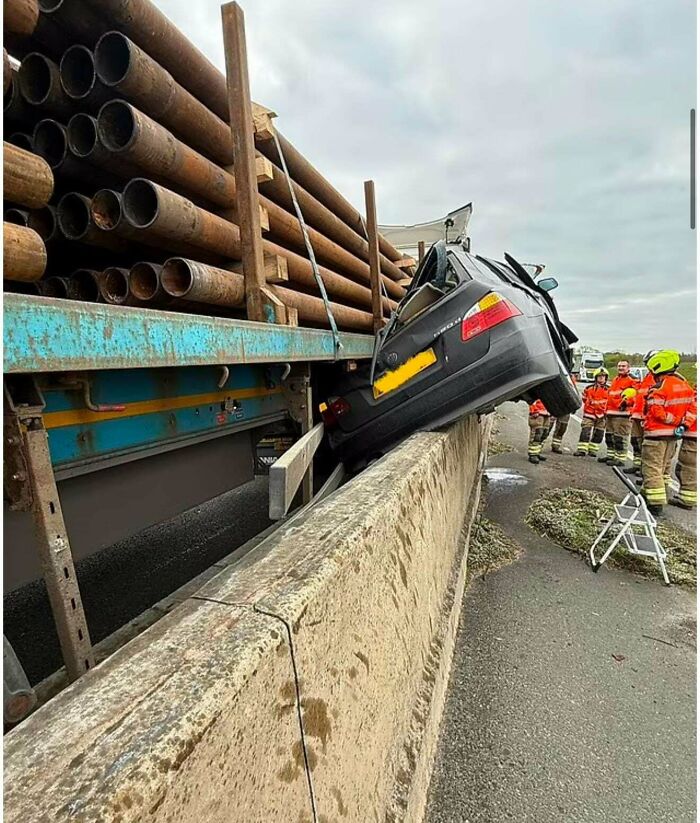 Car caught between concrete barrier and truck loaded with pipes, showing a serious parking fail and emergency responders nearby.