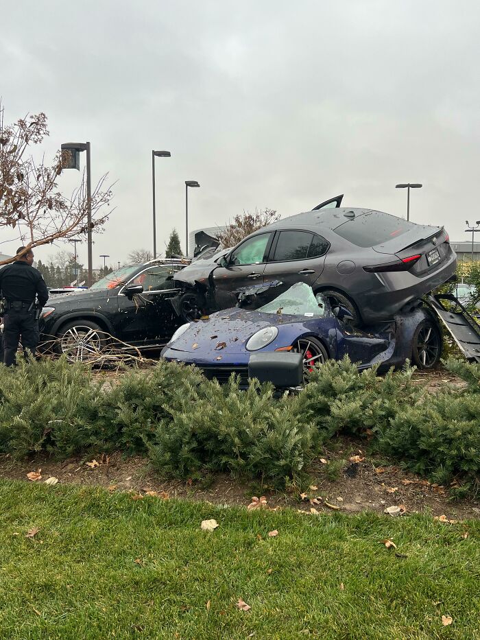 Police officer inspecting a severe parking fail with one car stacked on top of another in a parking lot near bushes.