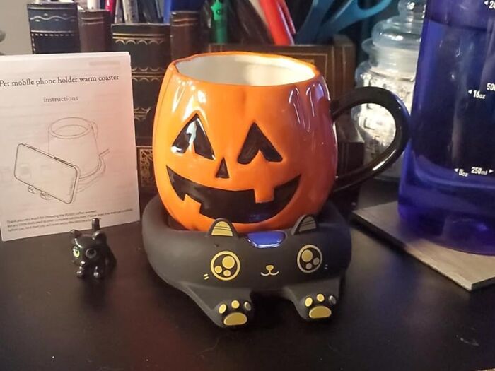 Halloween pumpkin mug sitting on a black cat-shaped mobile phone holder warmer among other desk items, showcasing useful decor.