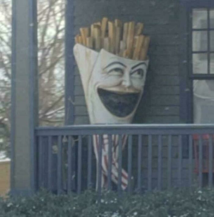 Creepy and unsettling statue of a smiling french fries container standing on a porch railing in dim light.