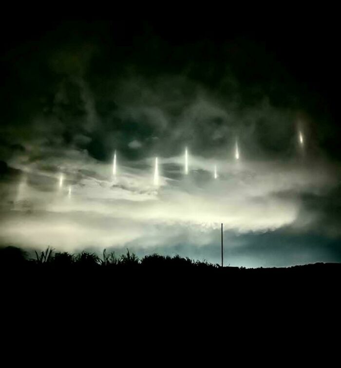 Unsettling night sky with eerie vertical light beams piercing through dark, creepy clouds over silhouetted landscape.