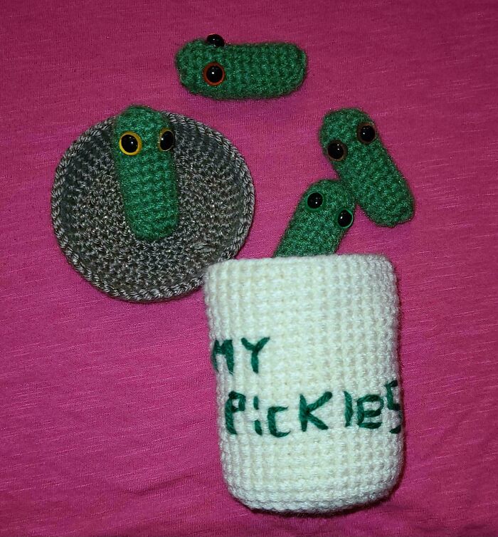 Crocheted green pickles with eyes, one on a coaster and three inside a white crocheted container on a pink background.
