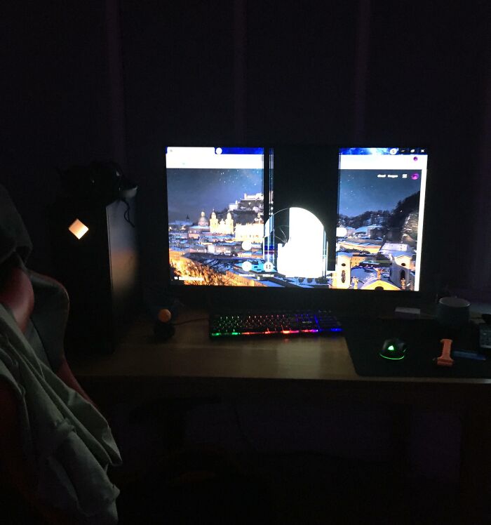 Dark room with a c*****d computer monitor showing a cityscape and a colorful backlit keyboard on the desk.