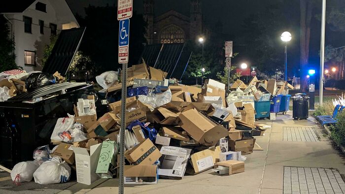 Overflowing trash bins with piles of cardboard boxes and consumer waste on a city sidewalk at night showing effects of consumerism.