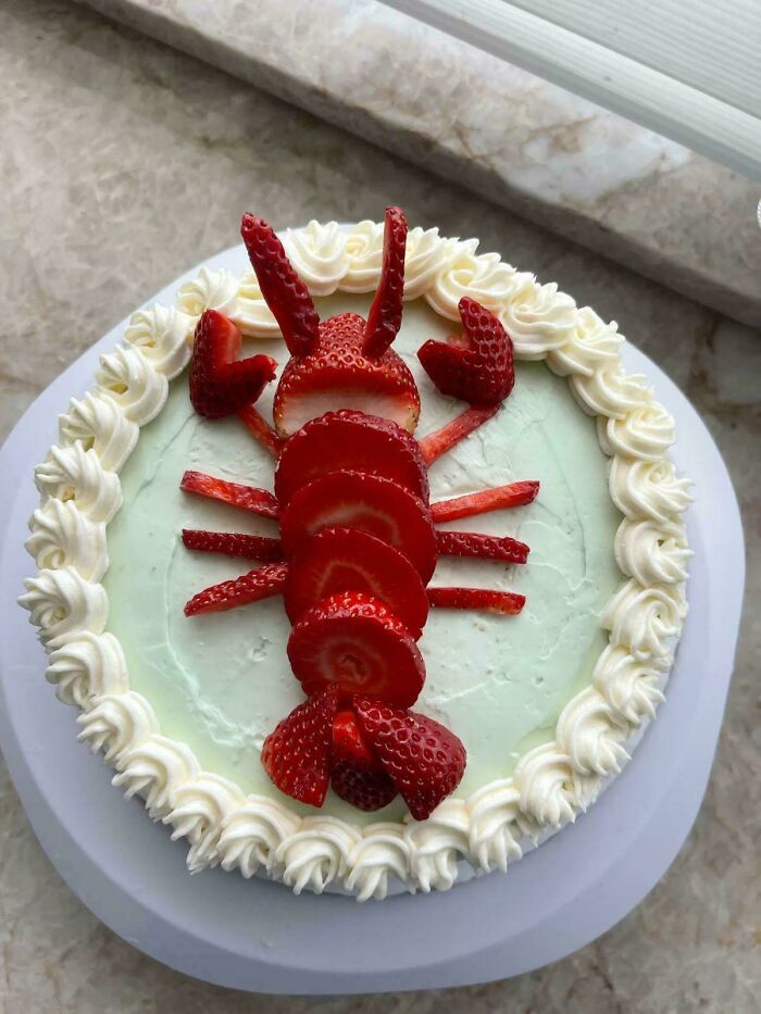 Round cake decorated with whipped cream and arranged sliced strawberries resembling a lobster, showcasing gorgeous baked goods.