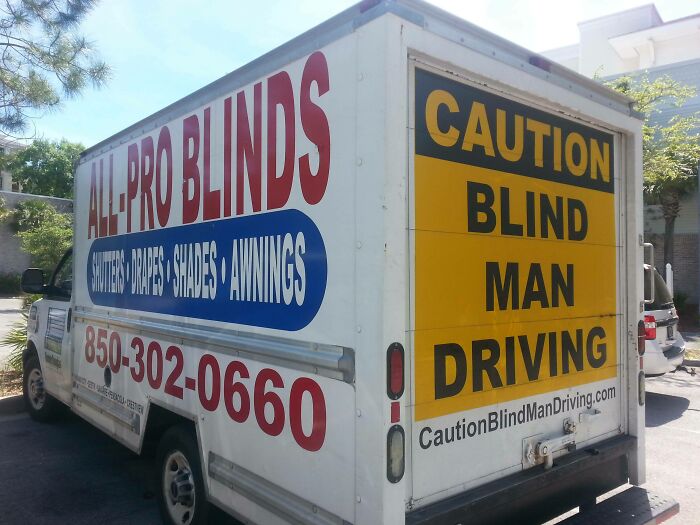 Delivery truck with a witty caution sign reading "Blind Man Driving" parked near trees and buildings in daylight.