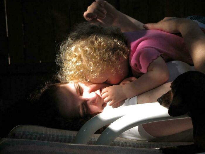 Child with curly hair leaning on adult lying on chair in soft light, creating an accidental renaissance photo moment.