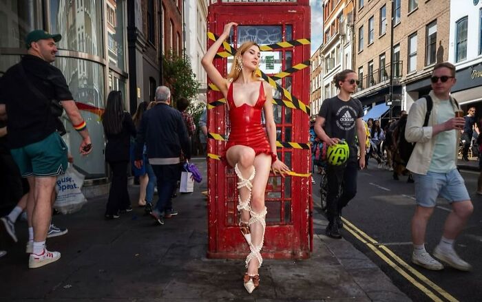 Woman in red dress poses wrapped in rope in front of a red phone booth, a powerful street photo revealing everyday life beauty.
