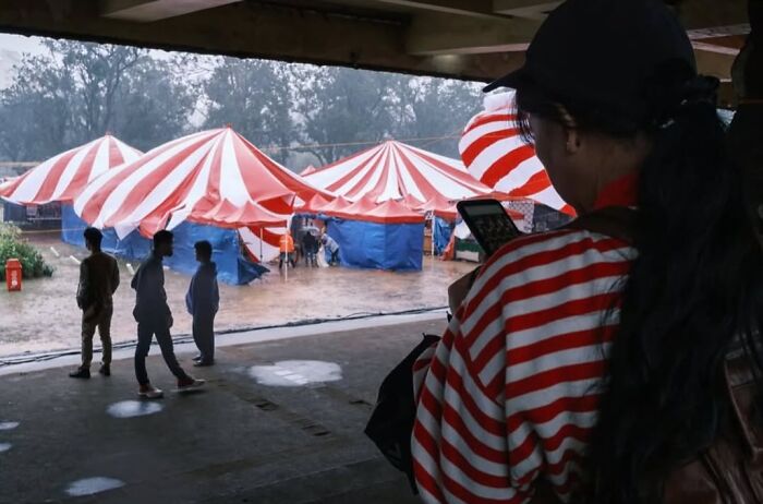 Woman in striped shirt using phone under shelter with people and red and white striped tents in rainy street scene.