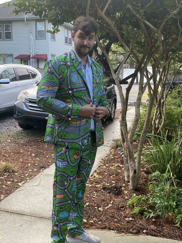 Man wearing a witty green suit patterned with colorful roads and buildings standing outside near trees and cars