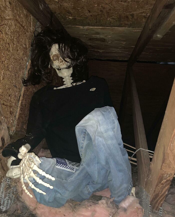 Skeleton dressed in black shirt and jeans with long hair wig sitting in attic, showcasing witty and humorous decoration.