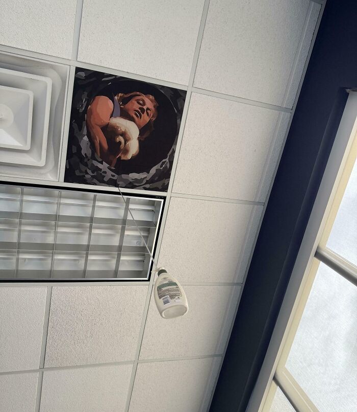 Ceiling with a missing panel replaced by a painting and a bottle hanging from a wire, showing wit in unexpected decor.