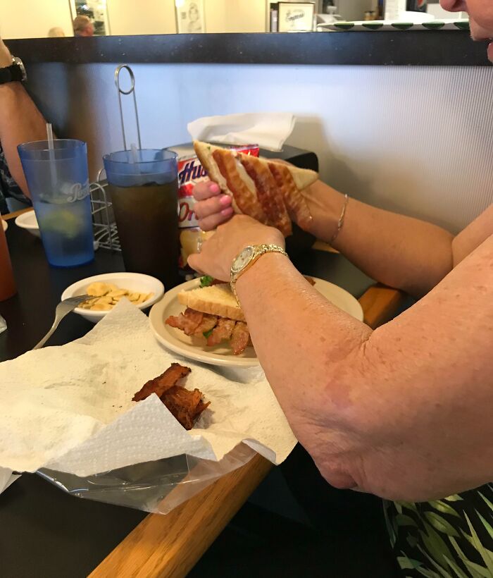 Person holding a large sandwich at a diner table with drinks, chips, and a watch on their wrist showing witty dining moment.