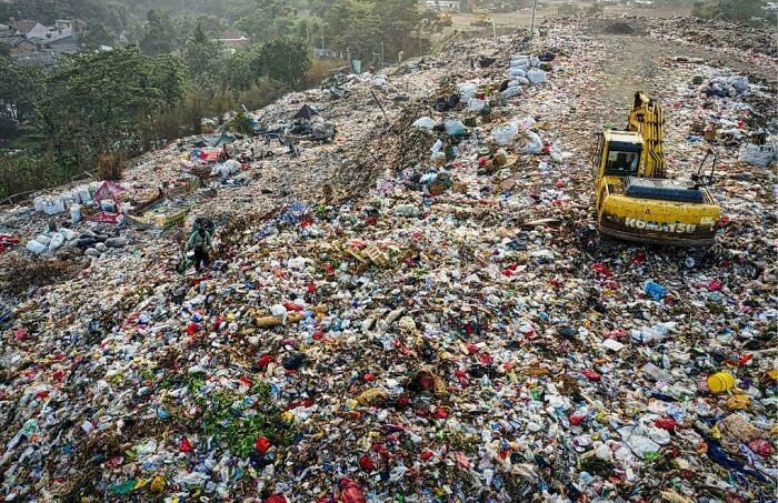 Massive urban hell landfill filled with scattered trash and a yellow excavator working amidst waste on a cloudy day.