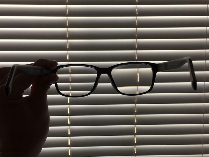 Hand holding eyeglasses in front of window blinds, illustrating visual comparisons with light and shadow patterns.