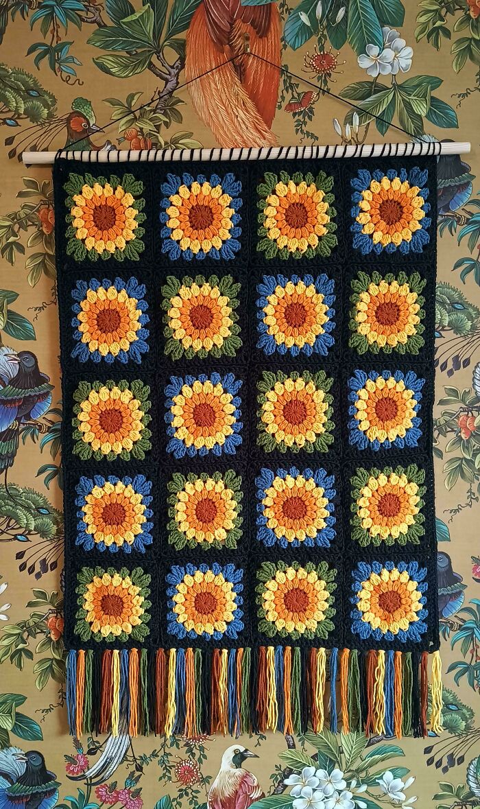 Crocheted wall hanging featuring multicolored sunflower squares in a grid pattern with fringe on a floral background