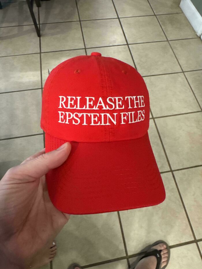 Red baseball cap with the phrase release the Epstein files held by a person on tiled floor, showcasing chaotic good attitude.