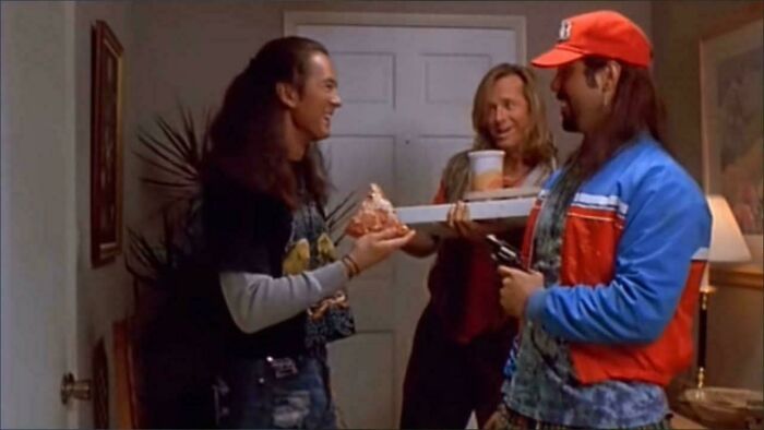 Three men in 90s style clothing sharing pizza inside a home, evoking 90s posts and nostalgic memories.