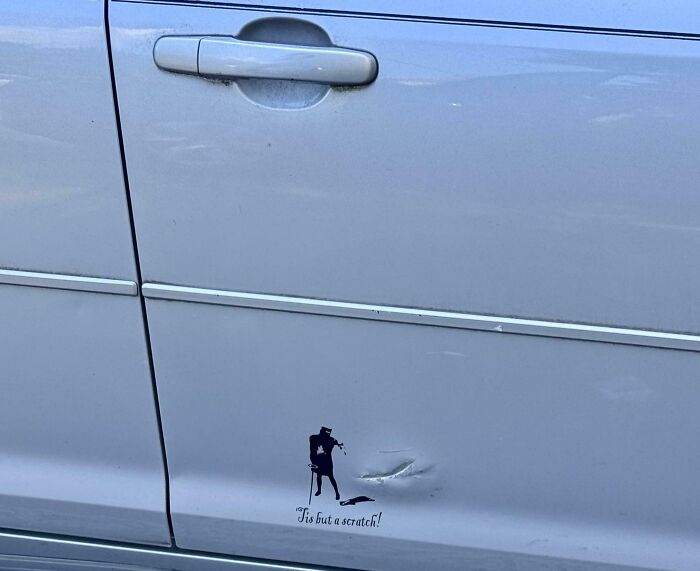 Silver car door with a dent and witty sticker saying Tis but a scratch featuring a silhouette in black.