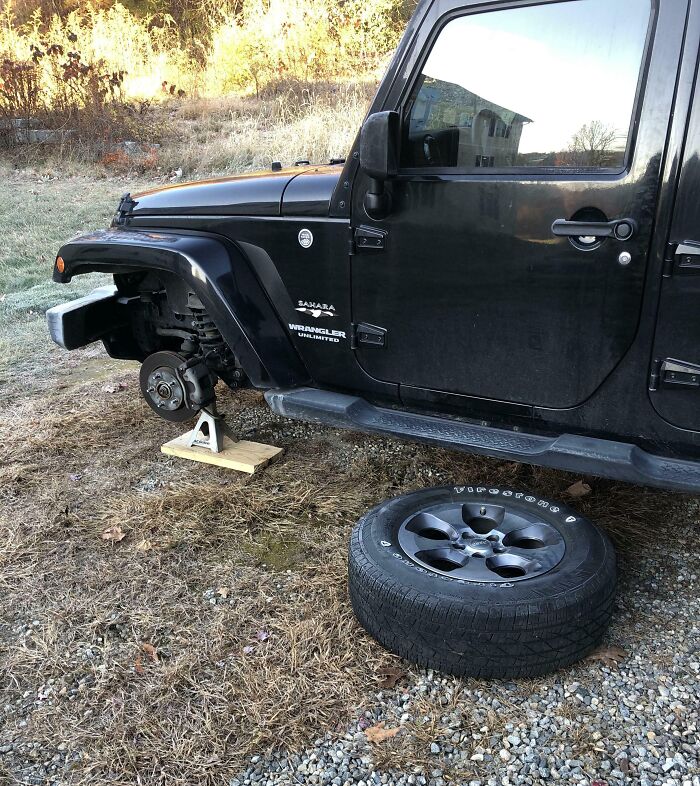 Black Jeep Wrangler without a front tire, supported by a jack stand on grass, showcasing witty car repair humor.