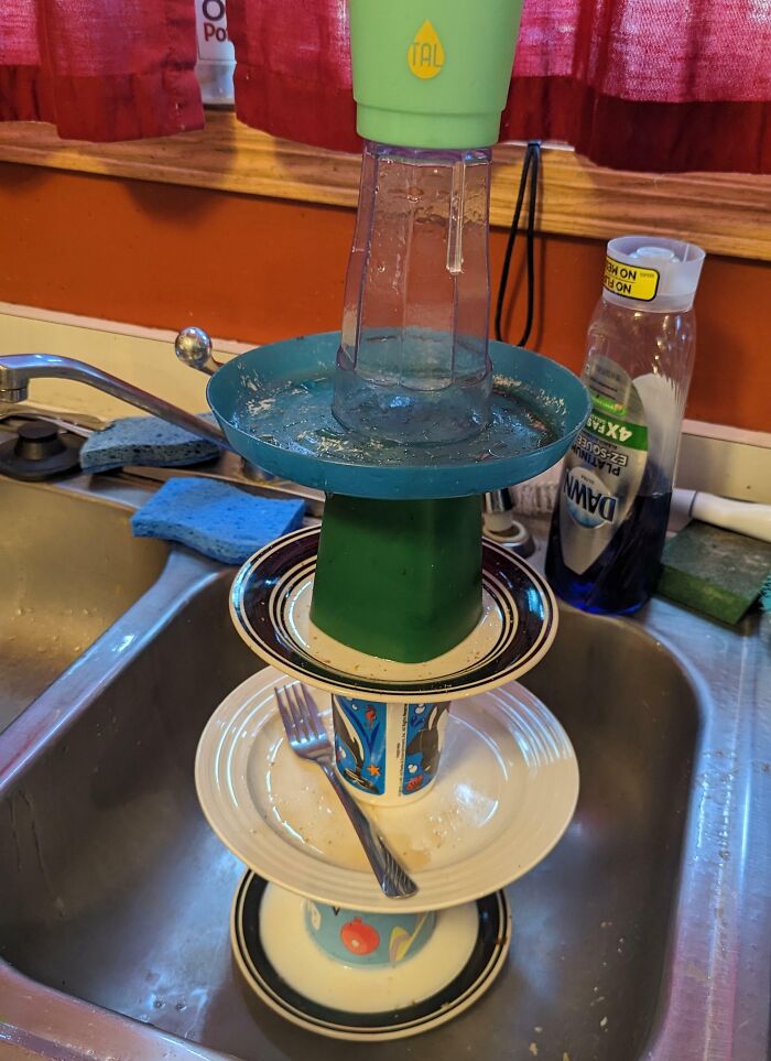 Stacked dishes and cups in a sink cleverly balanced to dry, showcasing witty kitchen organization and creativity.