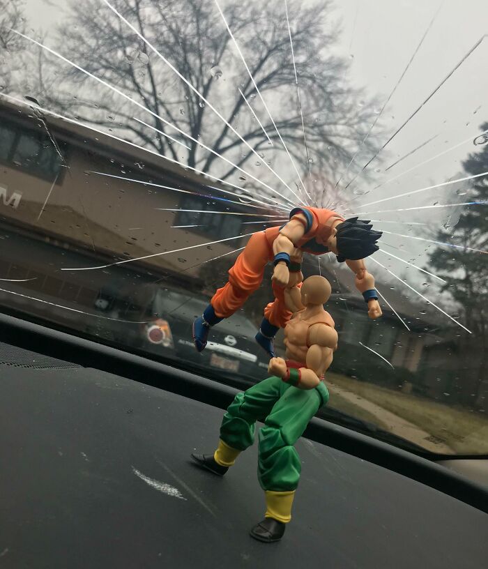 Two action figures staged in a witty fight scene on a c*****d car windshield with an urban background.