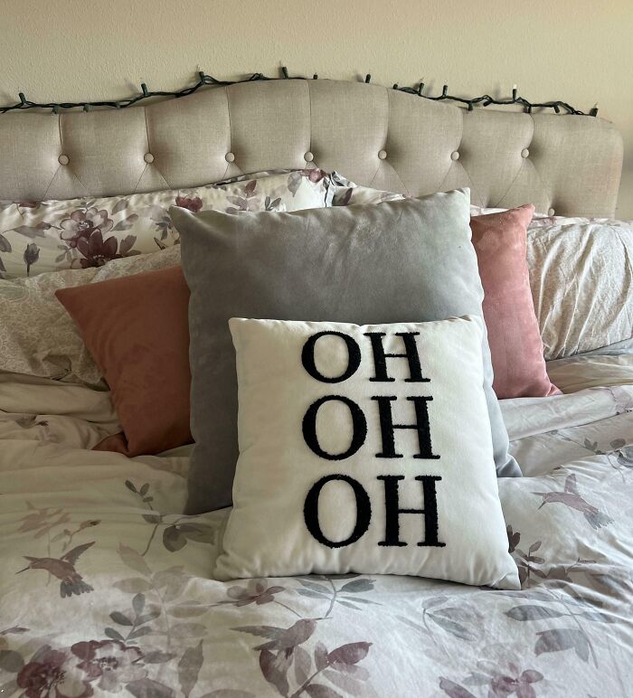 Decorative bed pillows arranged with a witty cushion featuring the word OH repeated three times on a floral bedspread.