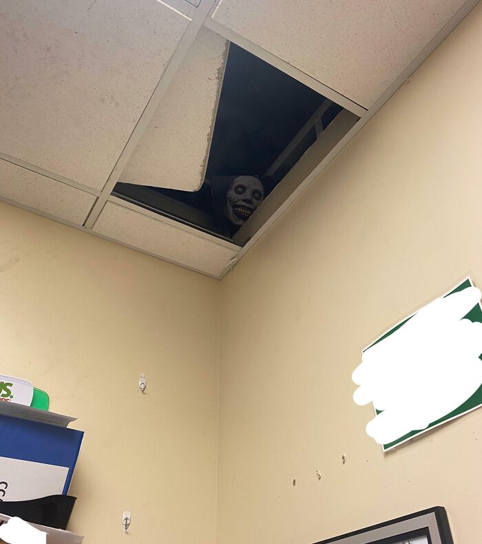 A creepy face peeking through a missing ceiling tile in an office, showcasing wit gone too far.