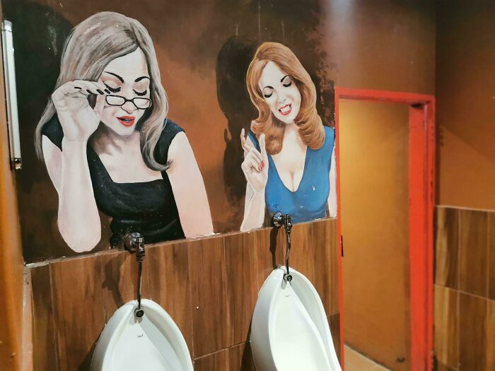 Bathroom wall art at a creative bar showing painted women interacting playfully above urinals in a unique global bar design.