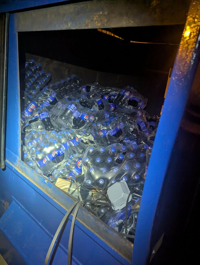 Dumpster diving treasures include dozens of discarded bottled drinks inside a blue metal dumpster at night.