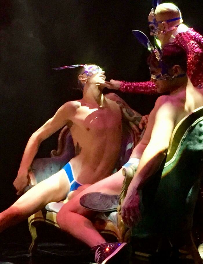 Three men in theatrical masks and minimal clothing pose dramatically on vintage chairs, capturing an accidental Renaissance photo.