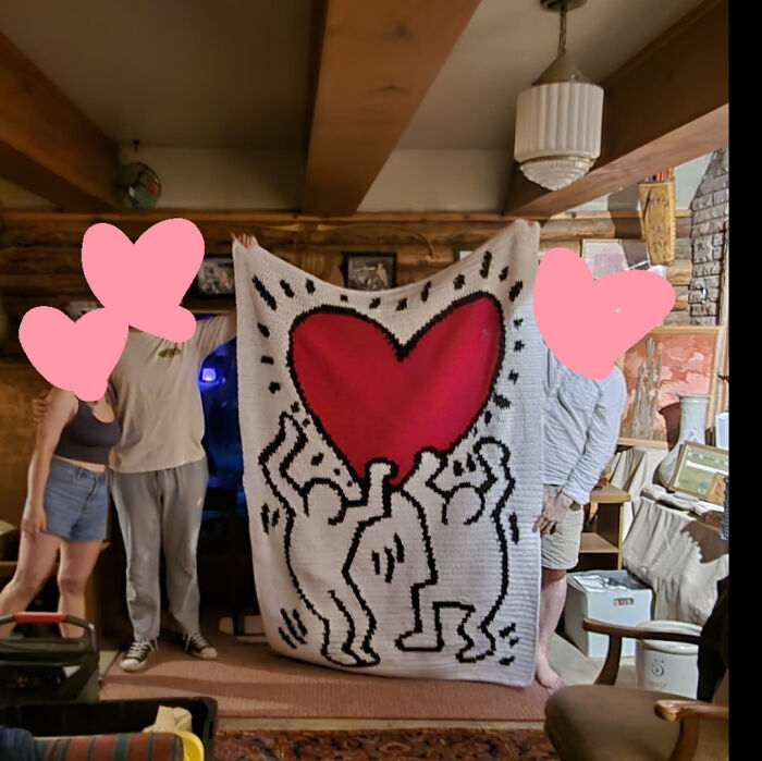 Three people proudly display a large crocheted blanket with a red heart and dancing figures design indoors.