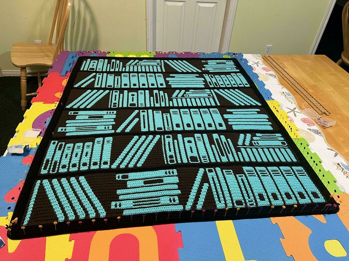 Crocheted blanket designed to look like a bookshelf, featuring intricate book patterns in blue and black yarn.