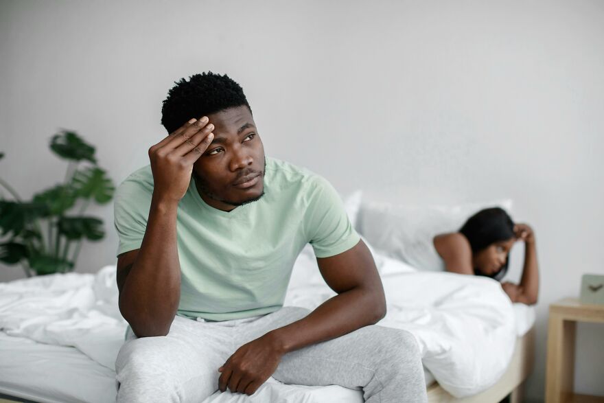 Man sitting on bed looking pensive and worried with woman lying in bed behind him, suggesting soul ties emotional struggle.