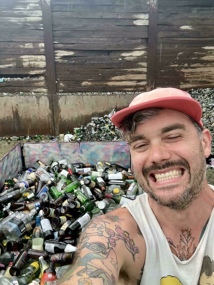 Man with tattoos and a pink cap smiling while standing next to a large bin filled with glass bottles, showing wholesome-kind neighbors.