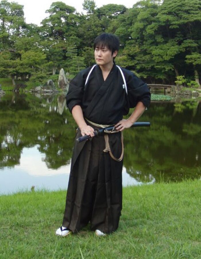 Man dressed as a samurai standing by a pond in a peaceful garden, illustrating superhumans walking among us.