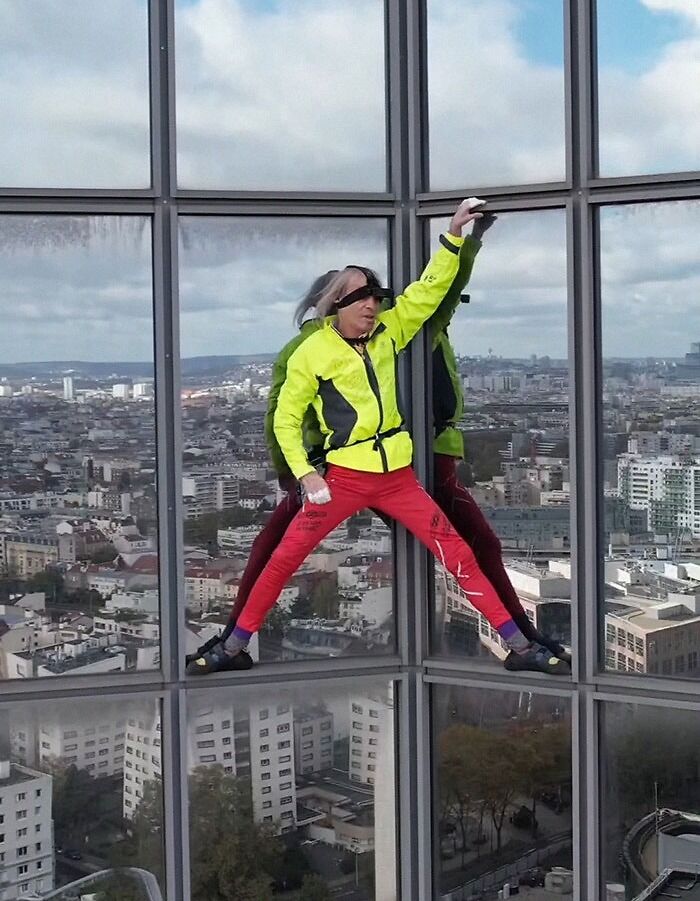Person dressed in bright clothes performing an extreme climbing act on a tall glass building, showcasing superhuman abilities.