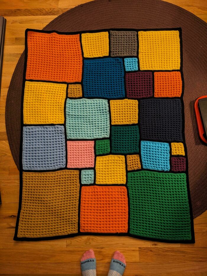 Colorful crocheted blanket made of various sized squares in orange, yellow, blue, green, and pink on wooden floor.
