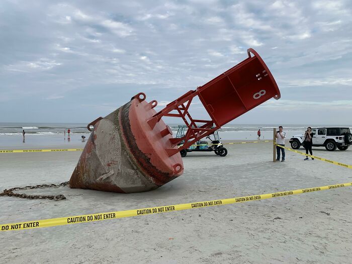 Large red buoy found at the beach surrounded by caution tape with people and vehicles in the background.