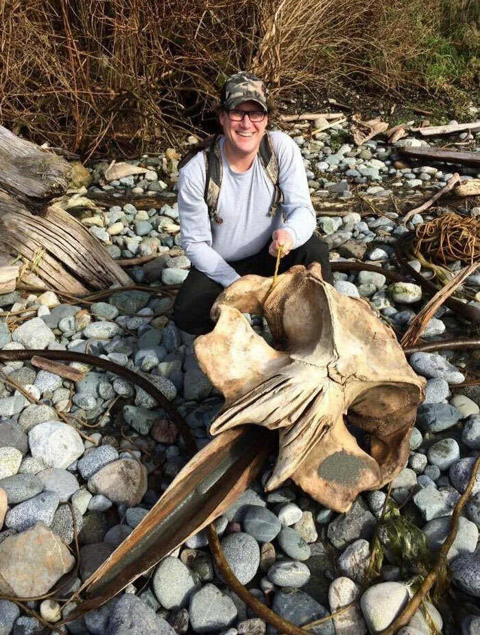 Person smiling on rocky beach holding large bizarre bone found at the beach among driftwood and stones