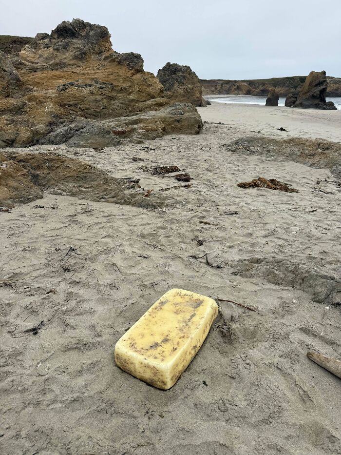 Large yellow block found buried in the sand near rocky formations on a beach with cloudy sky, an interesting beach discovery.