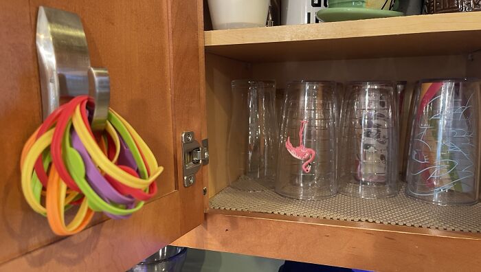 Colorful silicone bands organized on a cabinet hook next to neatly stored clear plastic cups inside a kitchen cabinet.
