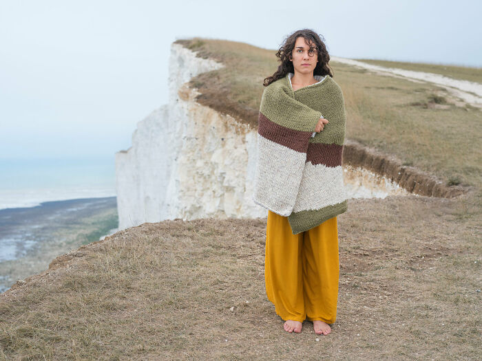 Woman wrapped in a blanket matching cliffside colors, blending with surroundings in a natural coastal landscape.