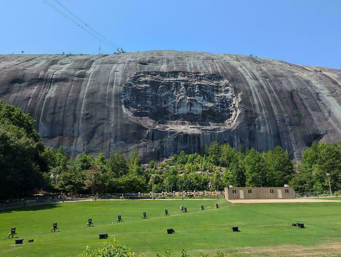 A carved mountain showing extremely large things, green grass, and trees under a clear blue sky, eerie.