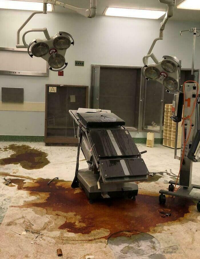 Abandoned operating room with a broken surgical table and large stain on the floor, creating a creepy and unsettling scene.