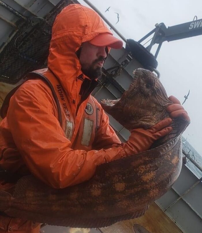 Man in orange rain gear holding a large unusual fish, capturing a creepy and unsettling moment on a rainy day at sea.