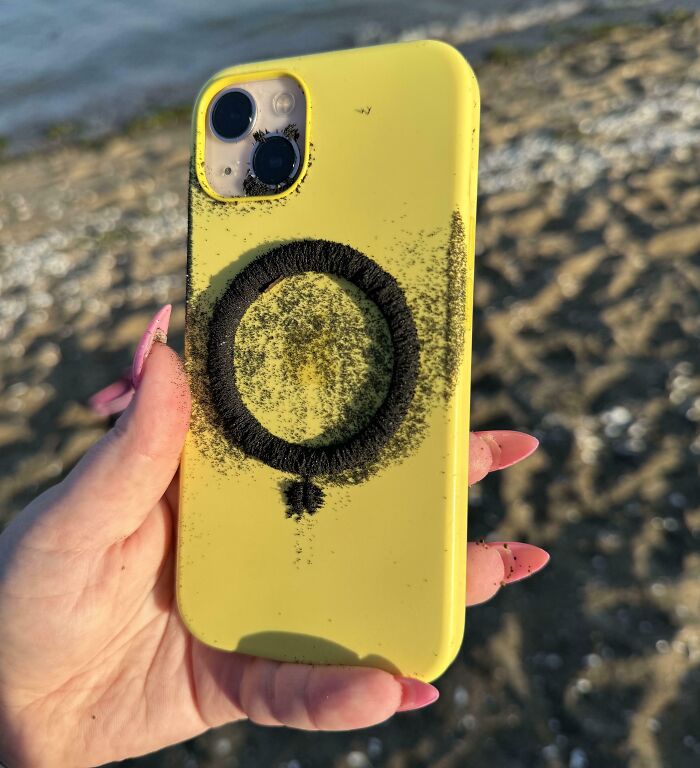 Yellow phone case with a black hair tie and sand forming an unusual pattern found at the beach by the shore.