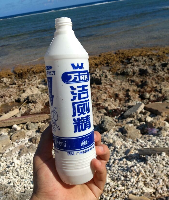 Hand holding a white bottle with blue Asian characters found at the beach with rocky shore and ocean background.