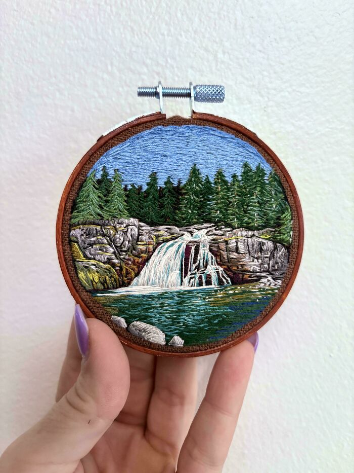 A stunning embroidery creation of a waterfall scene with pine trees and rocky cliffs, held in a hand.